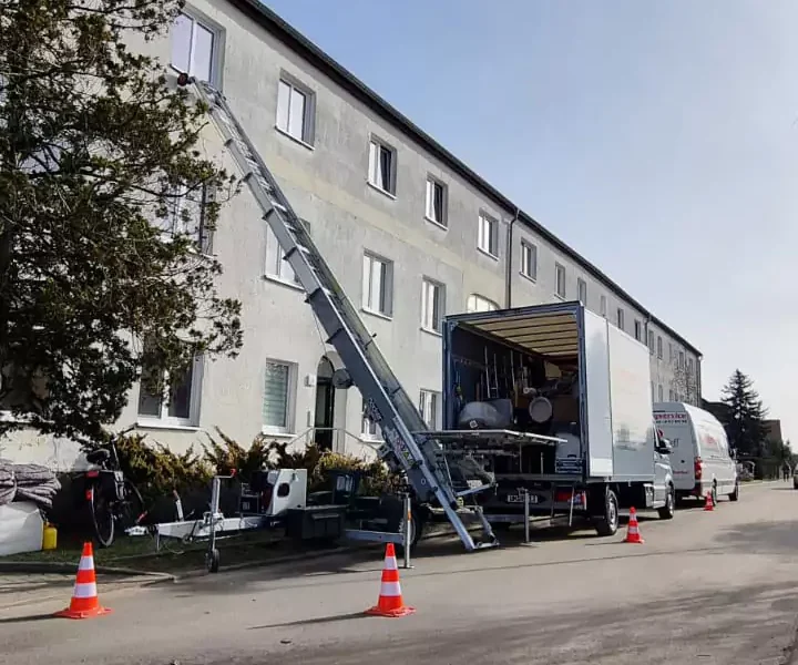 Umzugs-LKW und ein Sprinter stehen vor einem mehrstöckigen Wohnhaus an dem eine Leiter für den Möbellift aufgebaut wird